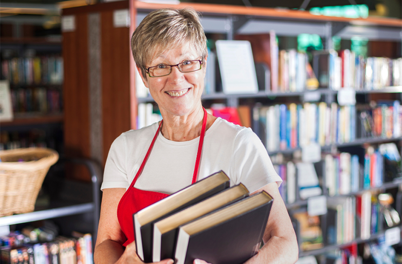 Adult white female with short blond hair holding books and standing in front of book shelves.