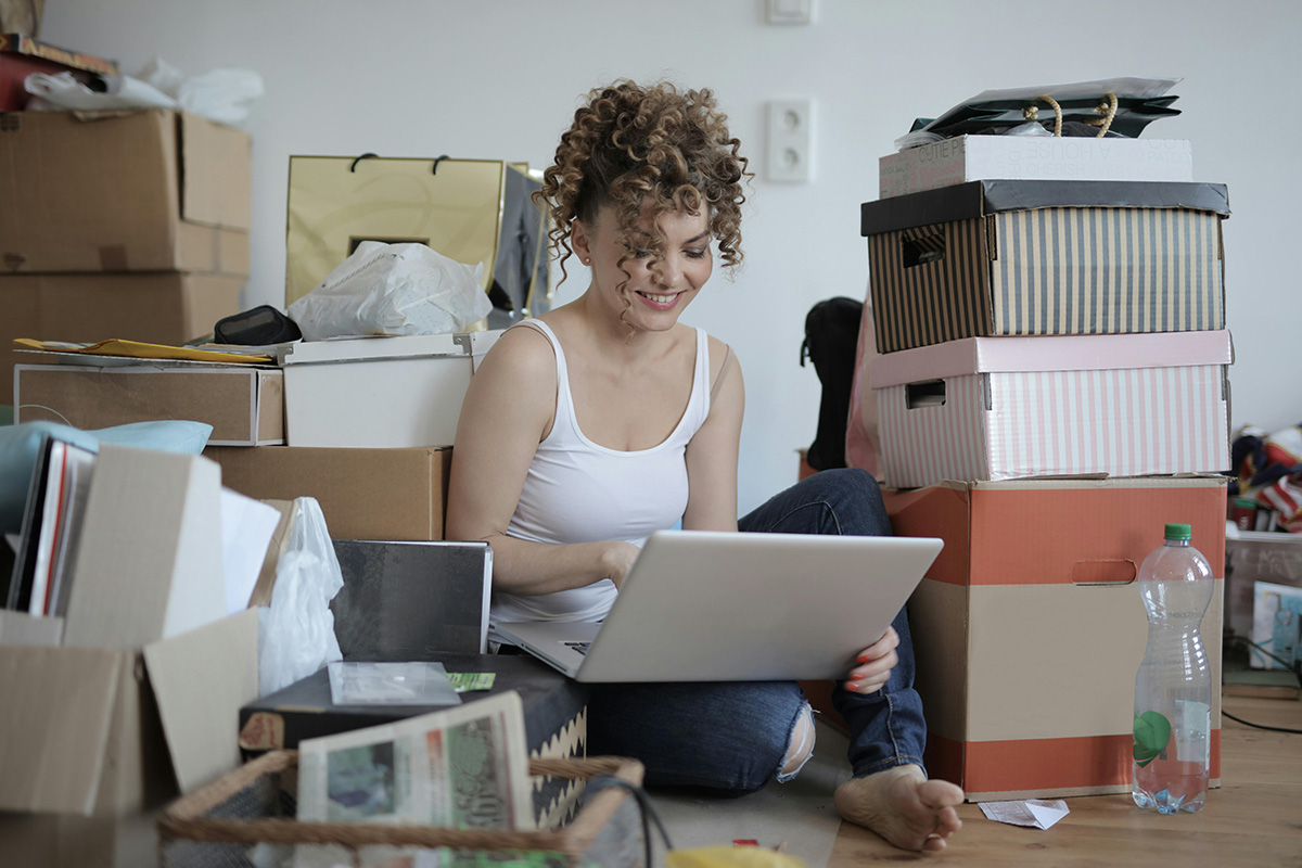 Young woman sitting on floor with laptop on lap amongst piles of books and stuff