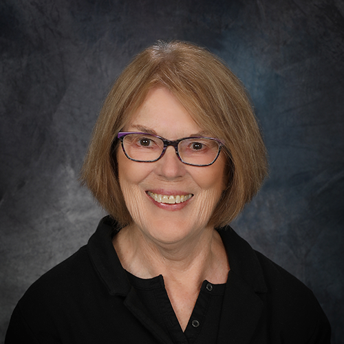 Older white female with purple framed glasses and straight short brown hair.