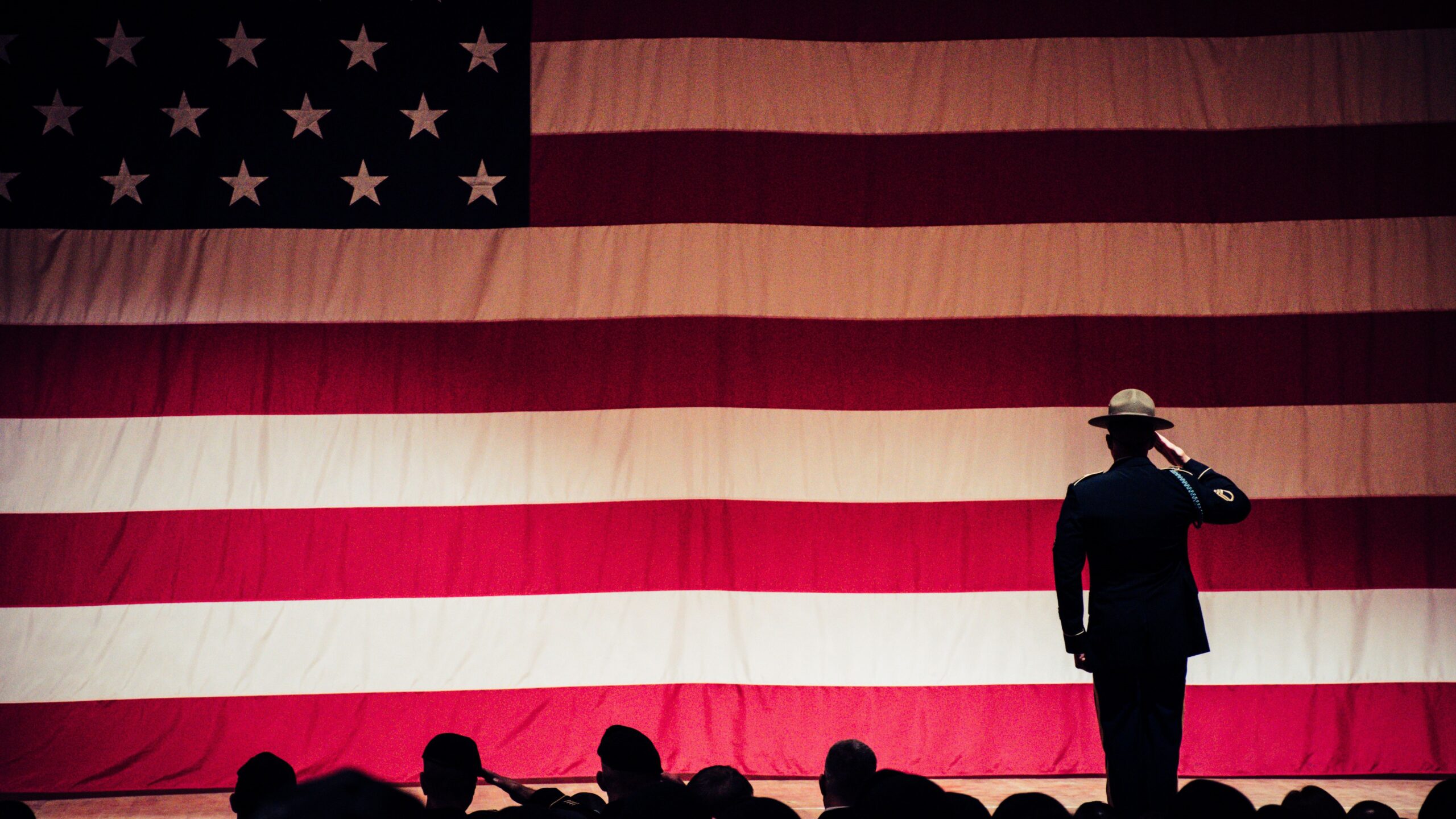 an American flag with a person in a military uniform standing at attention in front of the flag