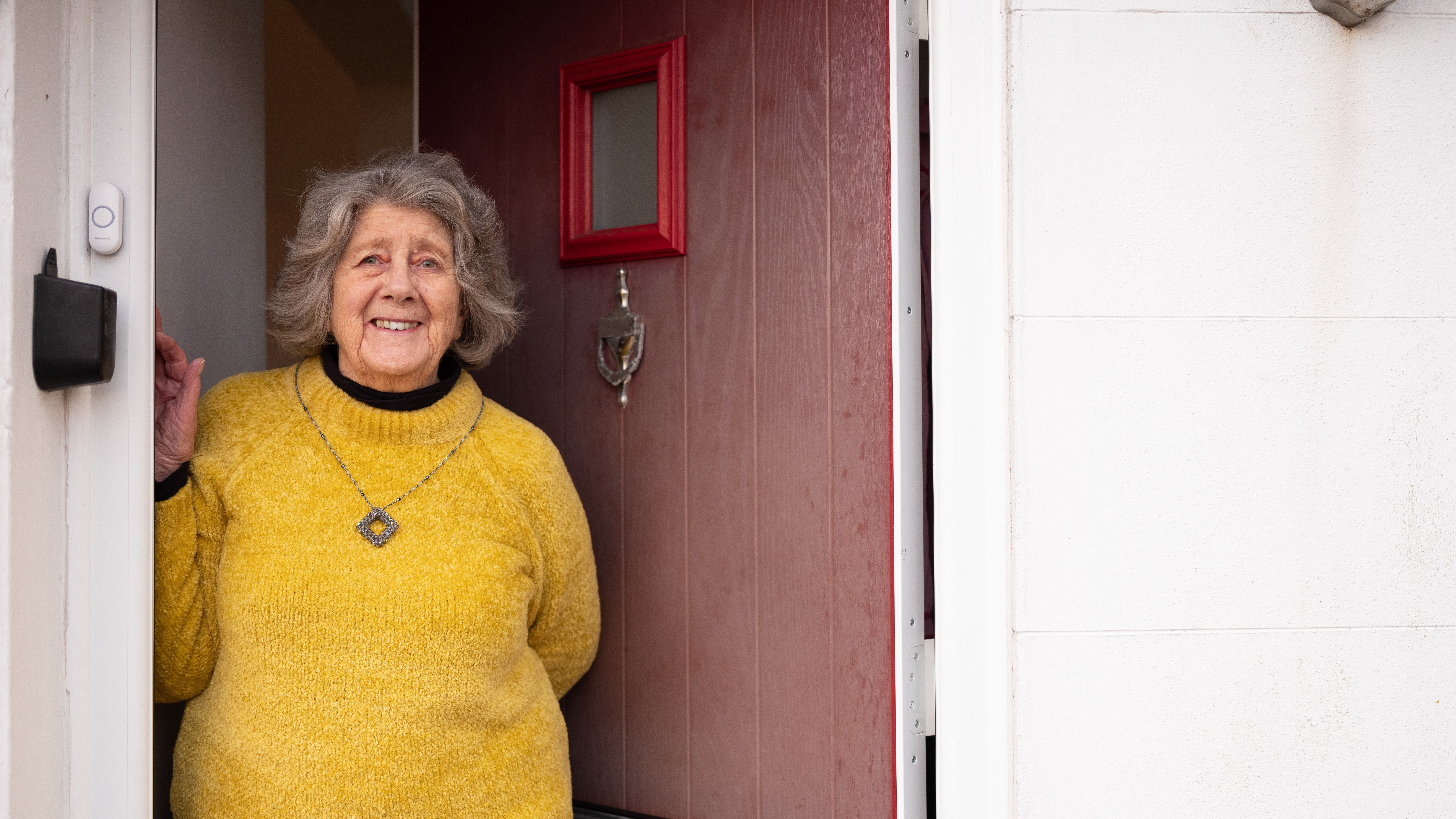 An older woman wearing a yellow sweater standing in the doorway of her home