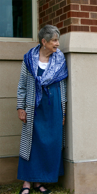 Older woman looking around brick building corner wearing long blue dress, blue and white striped cardigan, and blue figured scarf.